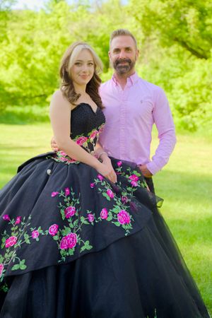 A ballgown featuring a black tulle skirt with bright pink floral embroidery details in an outdoor garden setting.