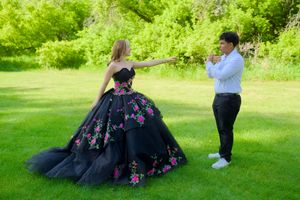 A couple poses in a garden setting with the woman wearing an elegant black floral ball gown.