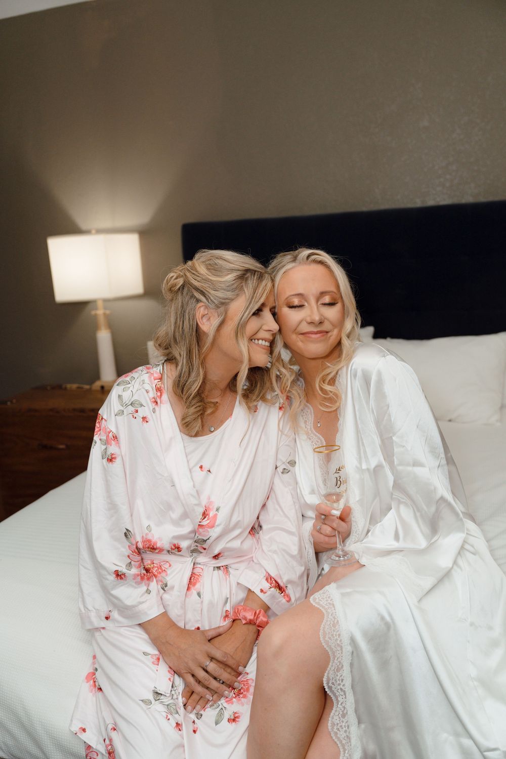 Two women in white floral and silk robes share a joyful moment together on a bed with a bedside lamp.