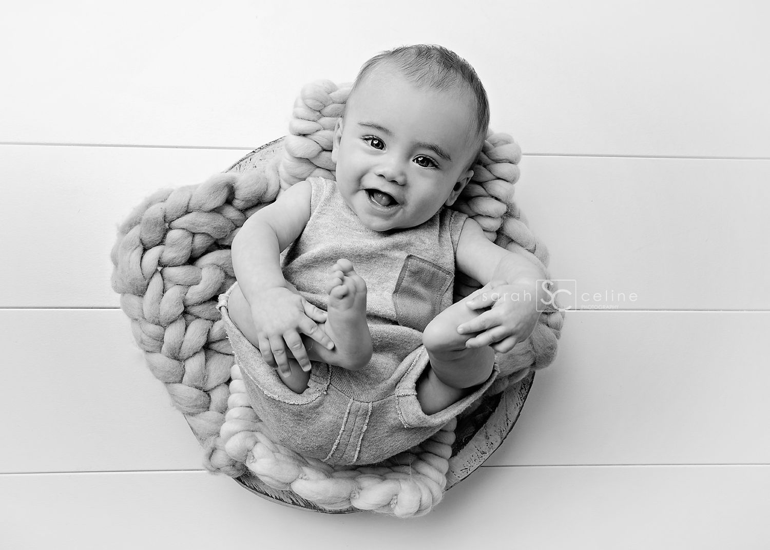 Black and white portrait of a smiling baby lying in a knitted basket on wooden floor background.