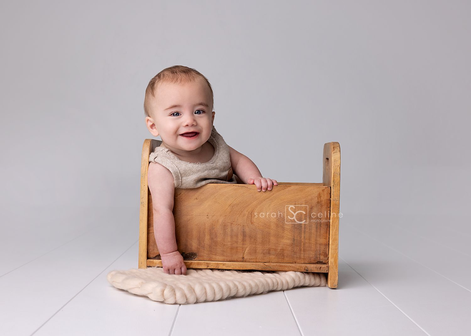 A smiling baby in a beige shirt sits inside a rustic wooden doll bed on a white floor.