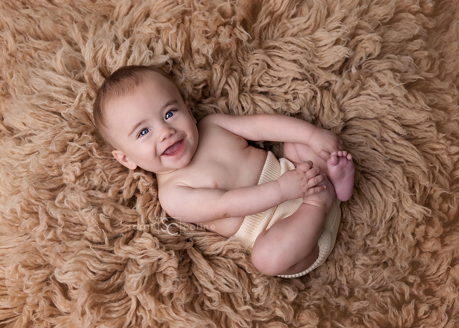 A smiling infant lying on fluffy beige carpet while holding their feet during a professional newborn photoshoot.