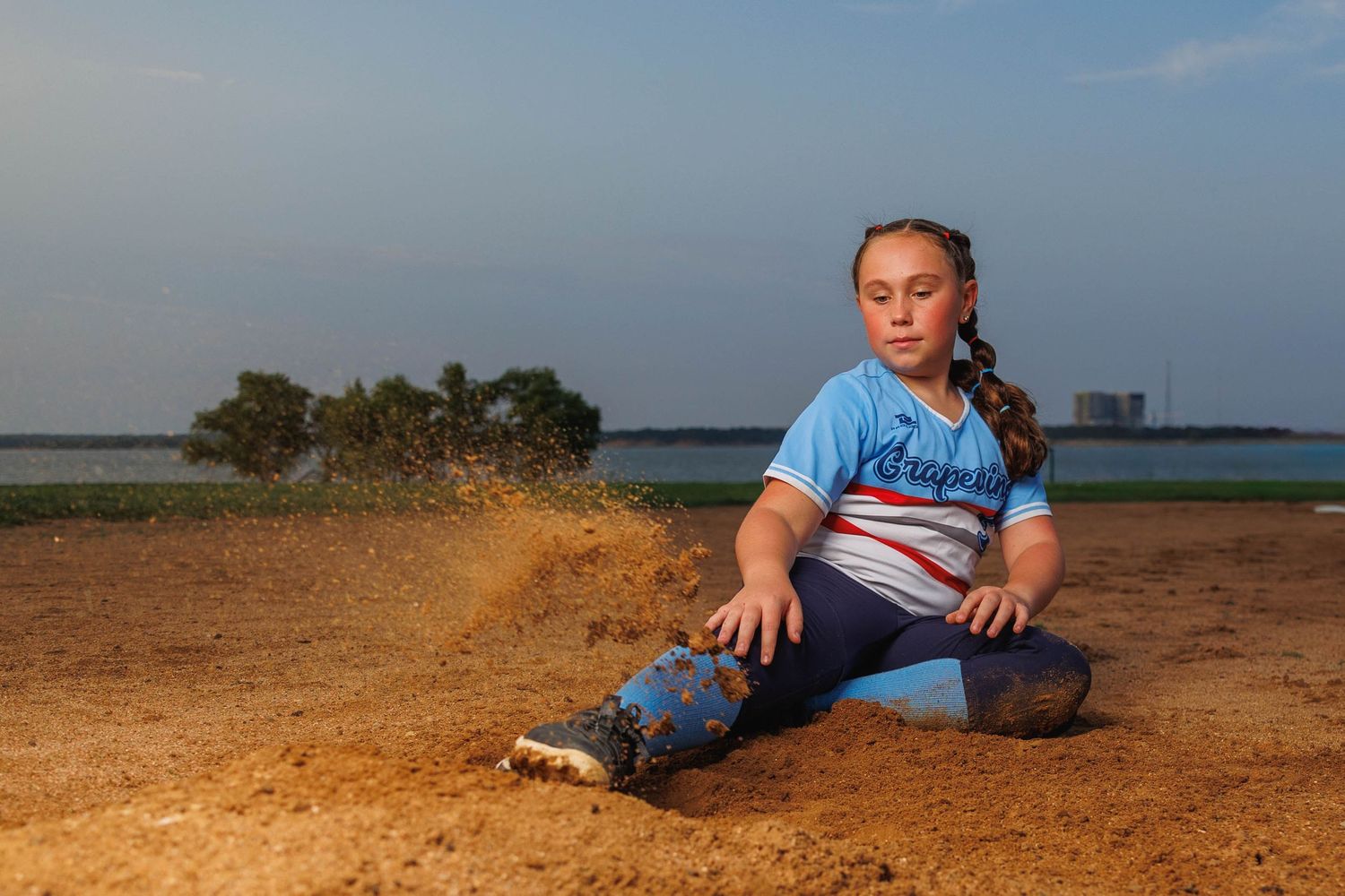Person sits on red dirt ground in blue t-shirt at dusk with distant trees and buildings on the horizon.