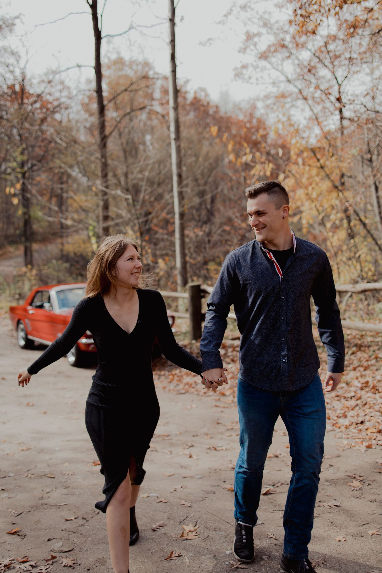 Couple walks hand in hand through an autumn forest path beside a vintage red truck.