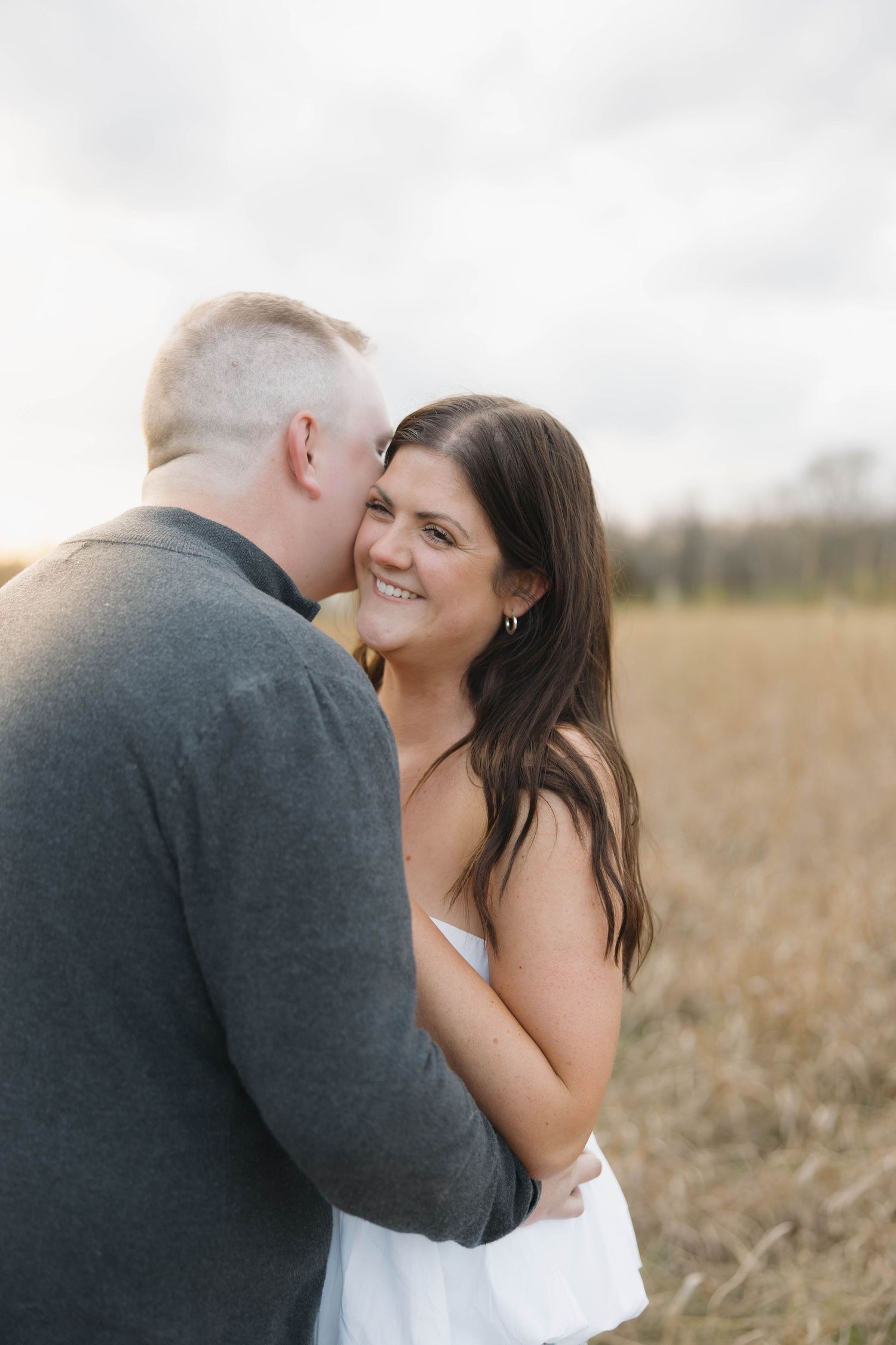 A couple embraces in a field of wheat during golden hour with cloudy skies in the background.