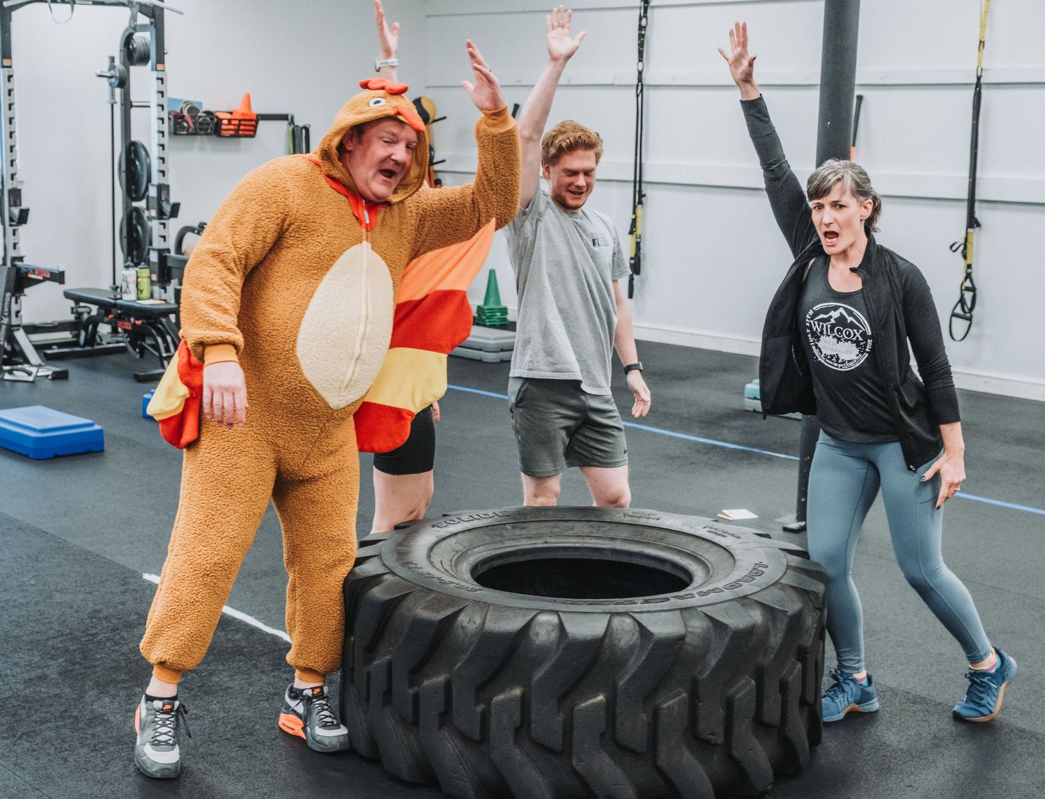 Energetic group of people exercising in a gym with a large tire, including someone wearing a brown animal costume.