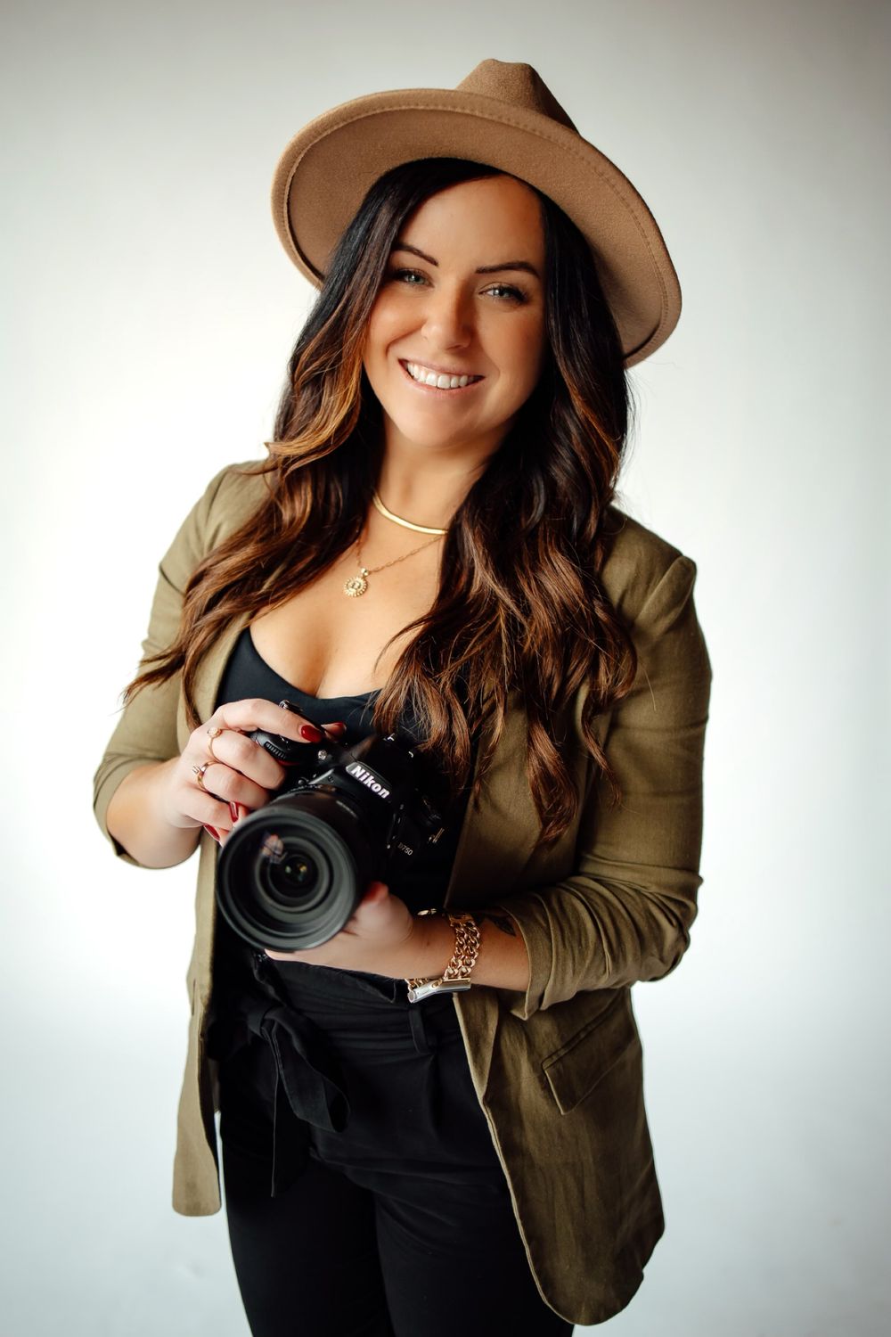 Professional female photographer smiling and holding a Nikon camera, wearing a tan hat and olive blazer.