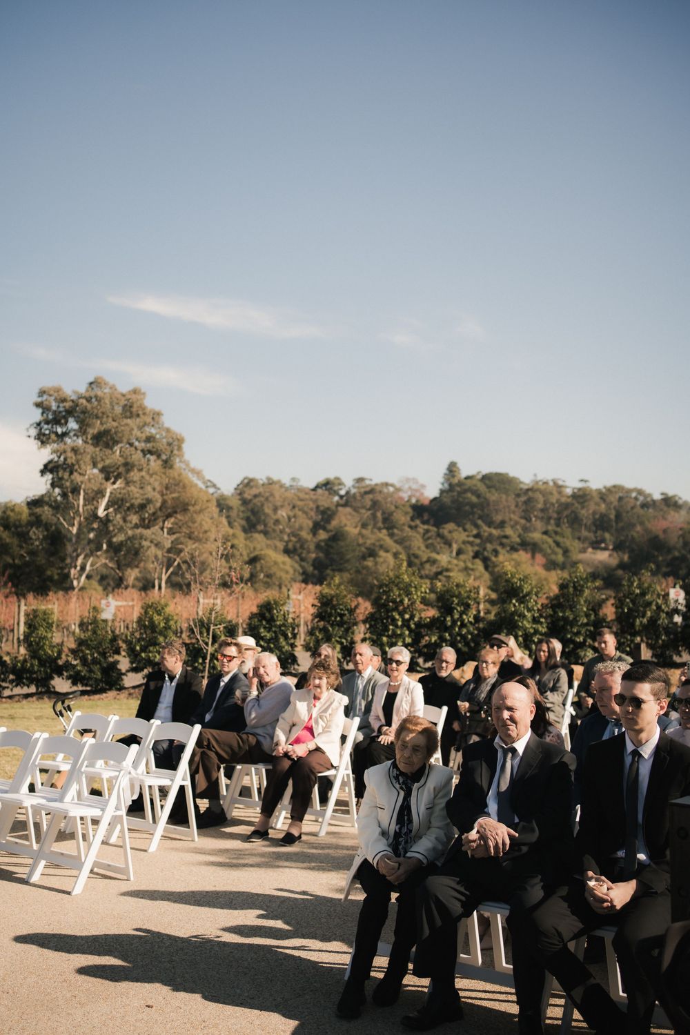 Wedding guests seated in white chairs at an outdoor ceremony venue with trees and blue sky in the background.