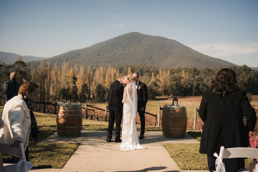 A wedding ceremony takes place at a scenic vineyard with mountains and wine barrels in the background during sunset.