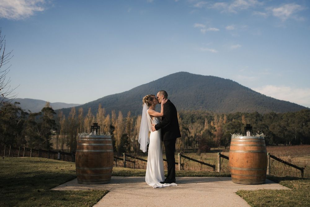 A couple stands on a pathway between wine barrels with mountain views at a rustic winery setting during sunset.