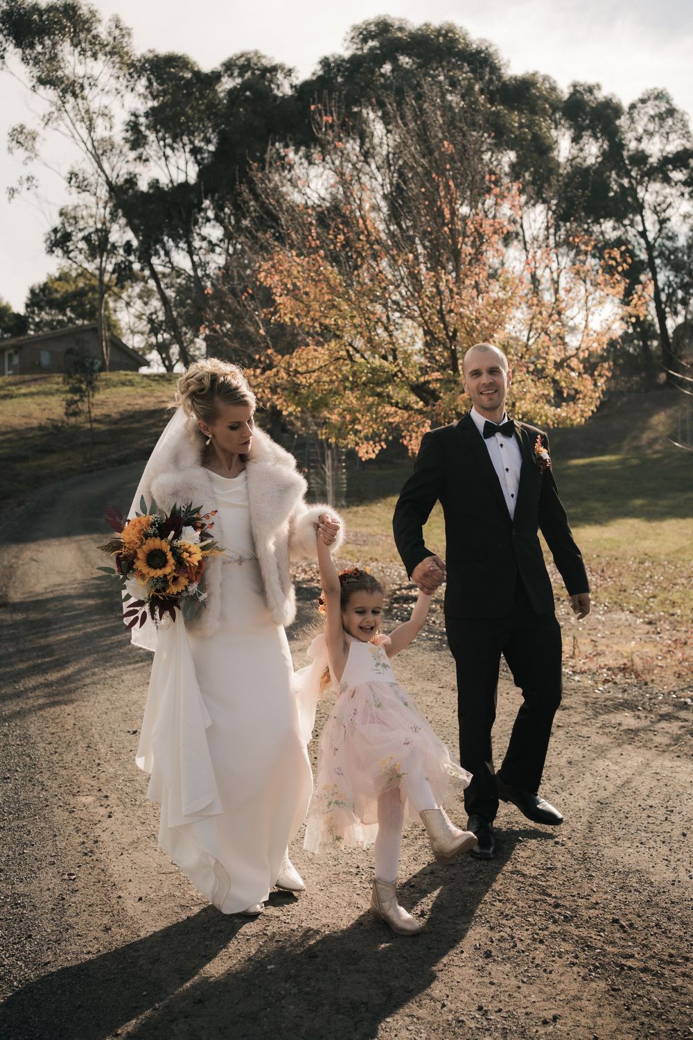 Wedding couple walks down path with flower girl while carrying sunflower bouquet against autumn trees.