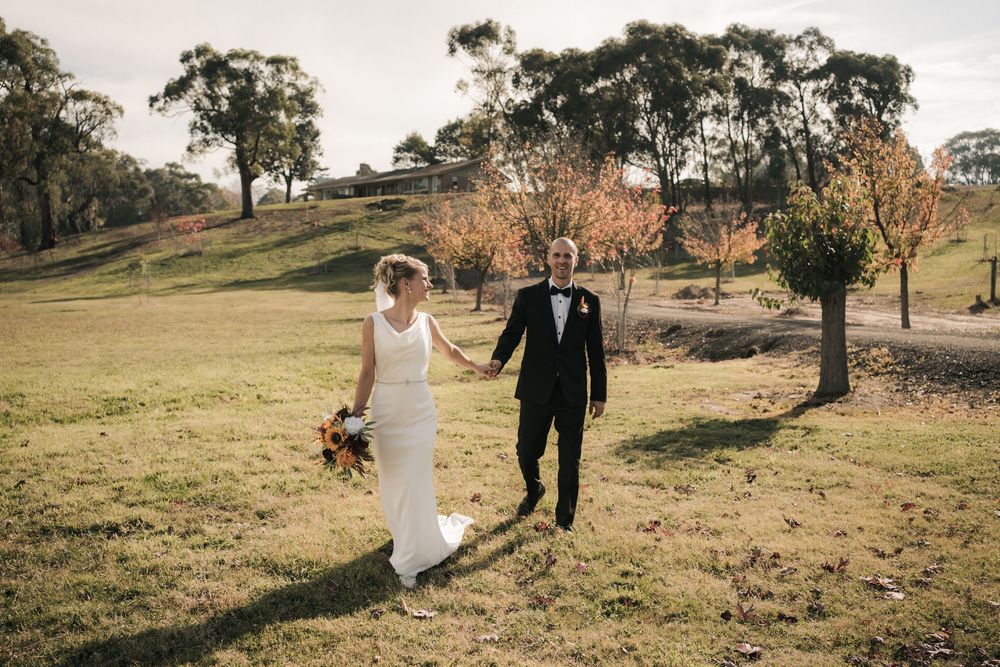 A couple dressed in wedding attire walks hand in hand through a scenic hillside with autumn trees in the golden sunlight.