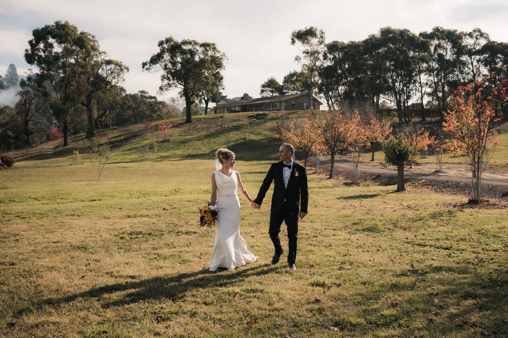 Two people in wedding attire walk hand in hand across a grassy hillside with tall eucalyptus trees in the background.