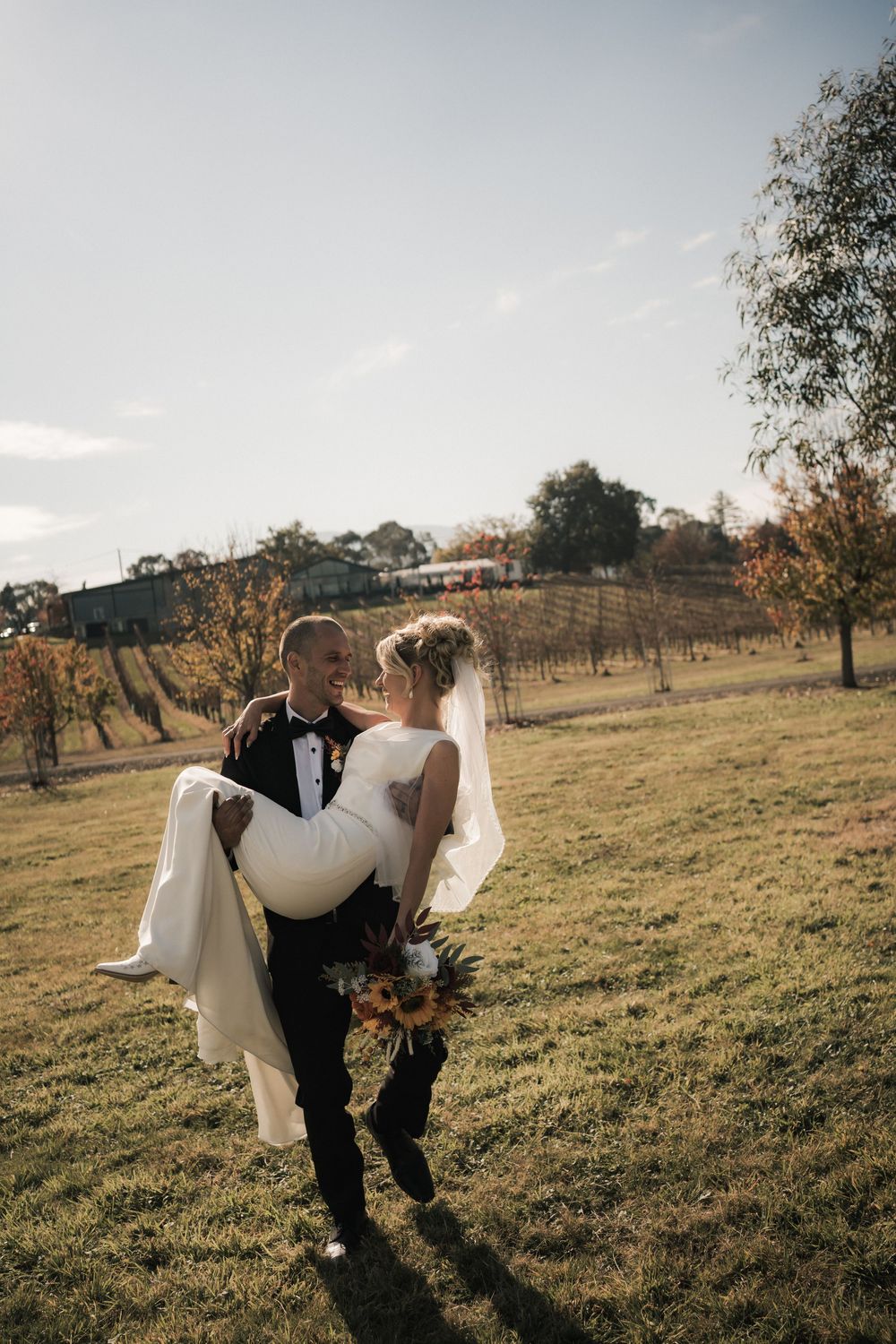 Wedding couple walking through a sunny field with golden grass and scattered trees.