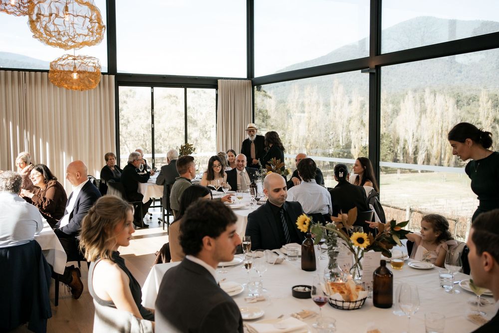 Wedding reception dinner setting with guests seated at tables in a modern venue with large windows.