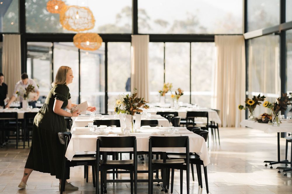 Event space with modern black chairs and white tablecloths being set up in a bright room with large windows.