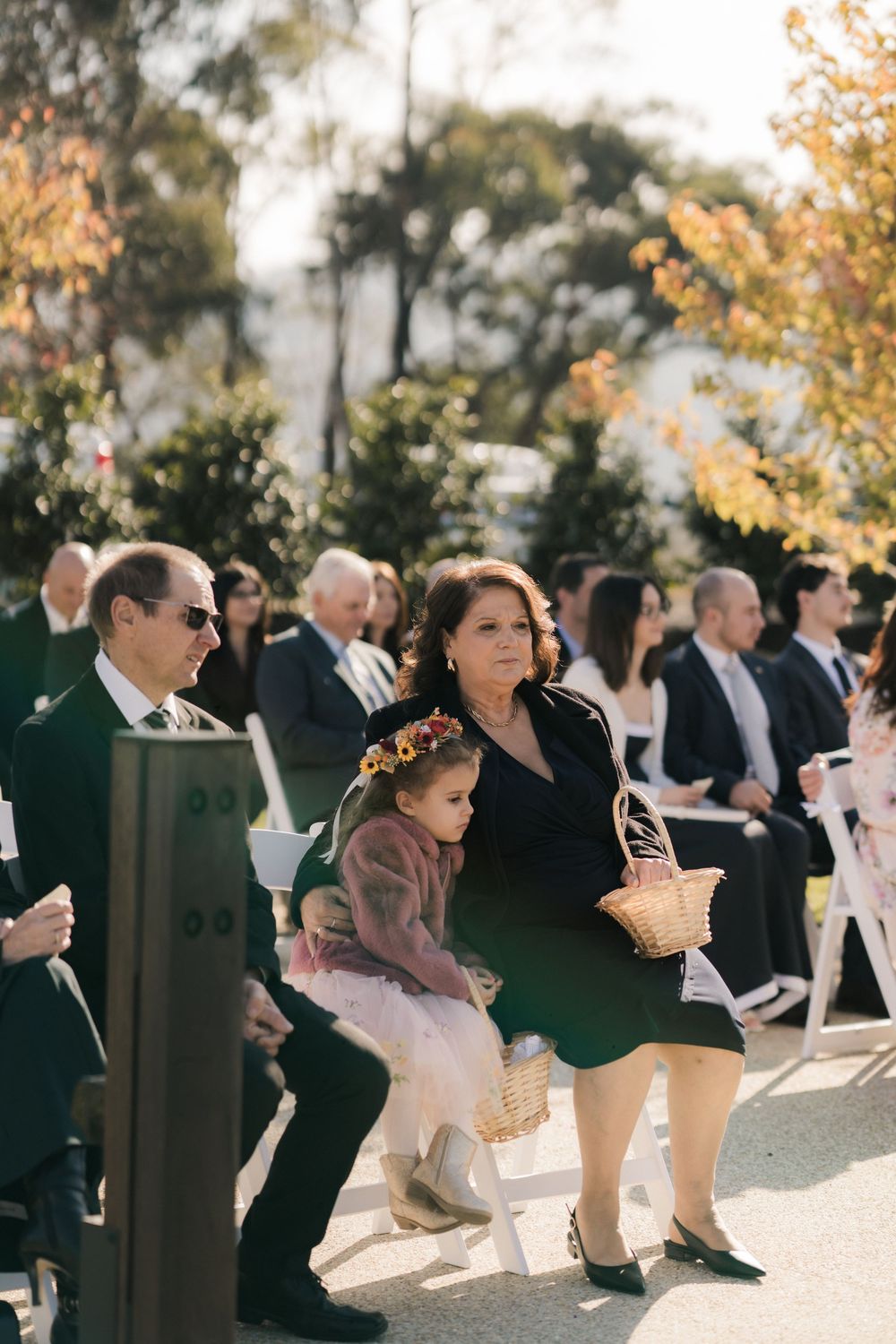 Wedding guests sit outdoors in white chairs during an autumn ceremony beneath golden-leafed trees.