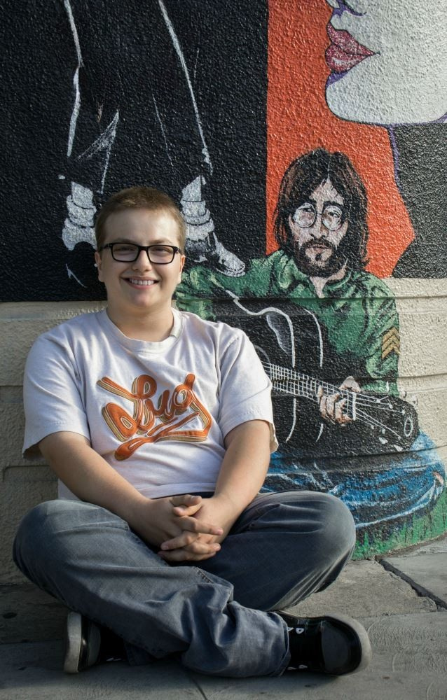 A young man sits by a wall of art while posing for a photo.