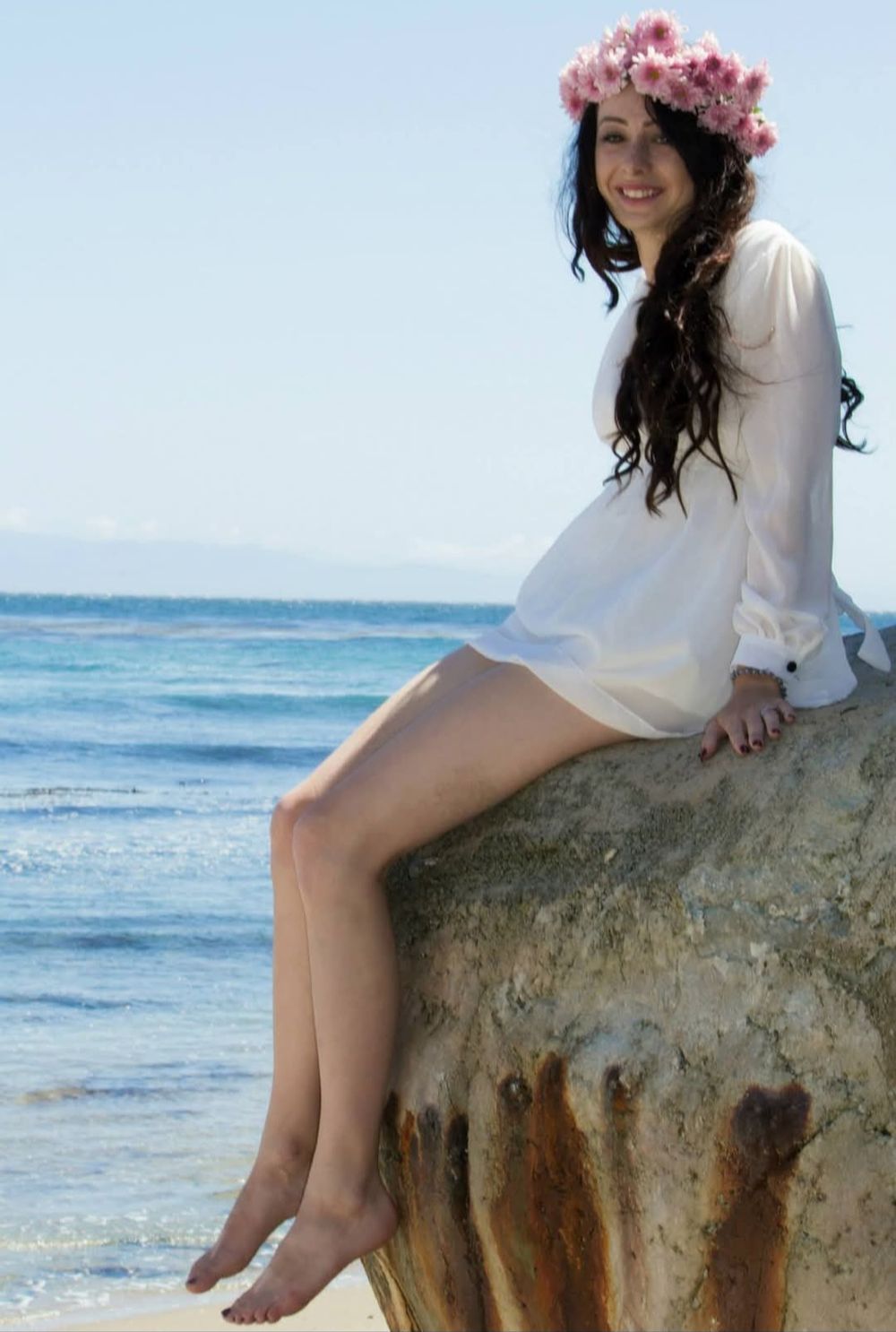 A young woman wearing a crown of flowers poses for a photo while sitting on top of a rock near the ocean.