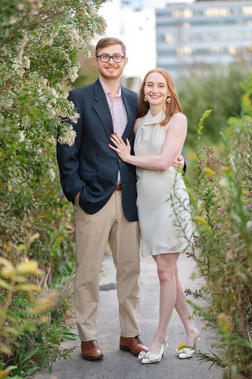 Smiling couple embracing among flowering bushes in urban garden, woman in white dress, man in navy blazer.