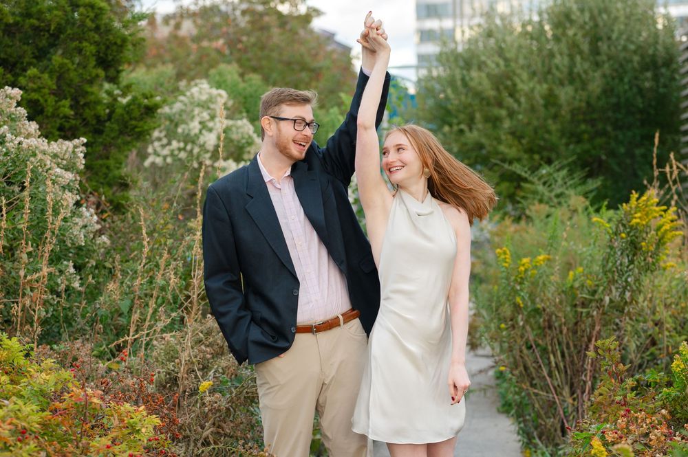 A joyful couple dances on a garden path, the man twirling the woman in a white dress amid lush greenery.