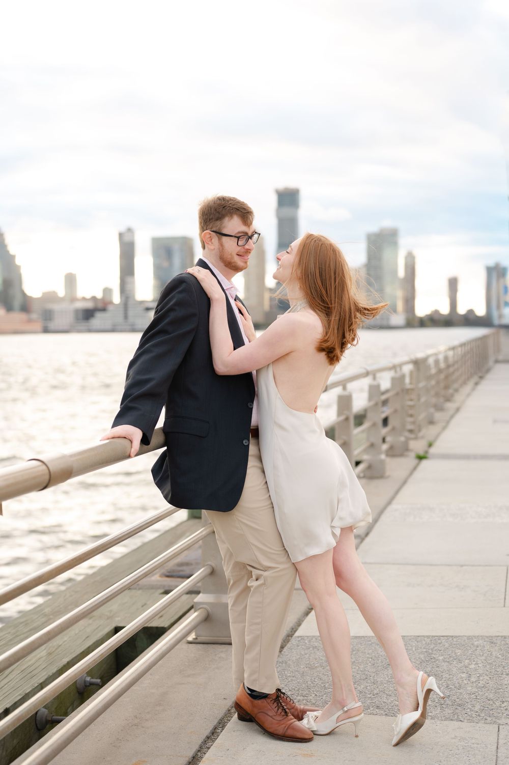 Couple embracing on a waterfront promenade with a city skyline backdrop, woman in white dress and man in navy blazer.