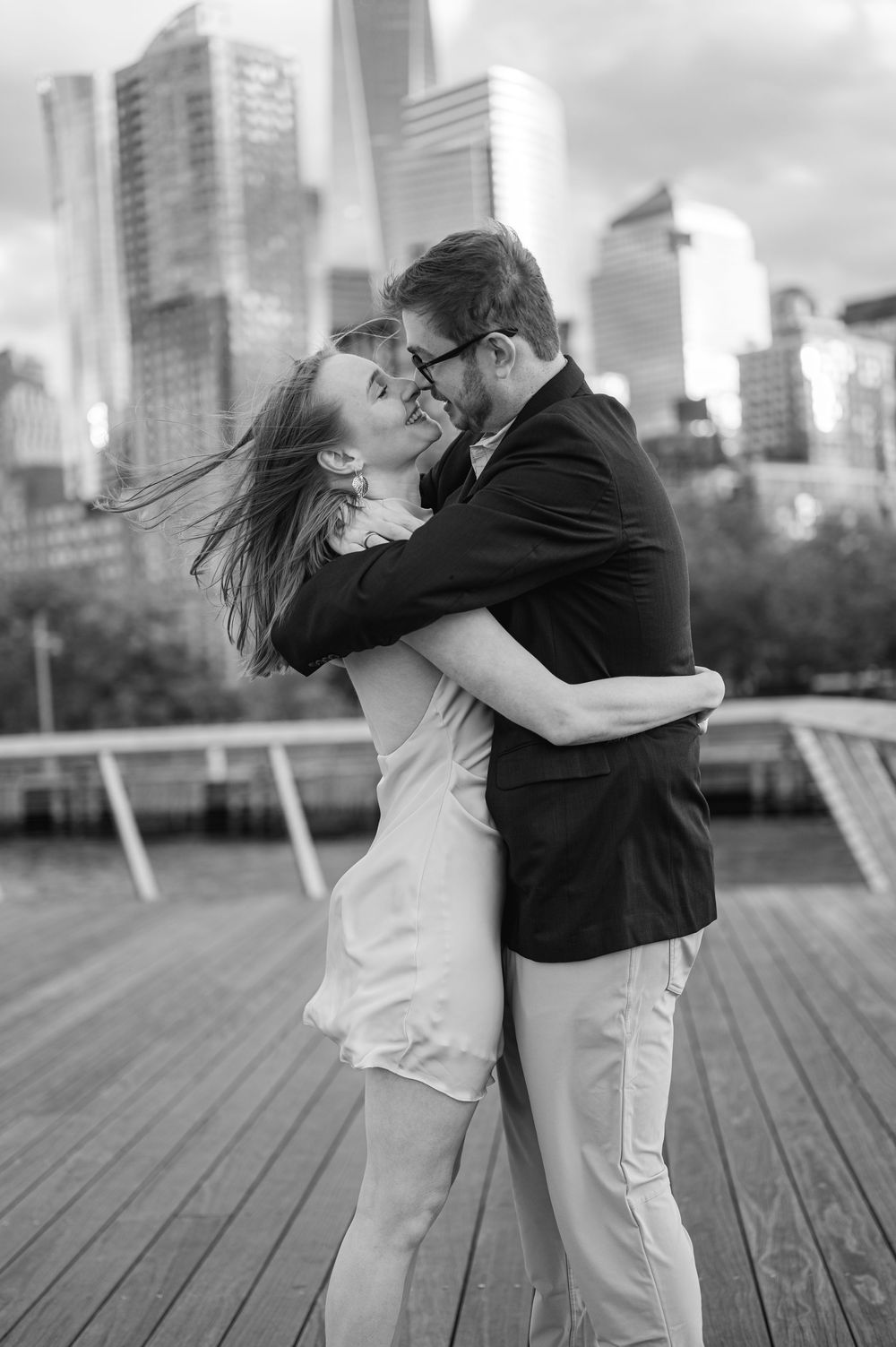 Black and white photo of a couple kissing on a waterfront dock with city skyline behind them.