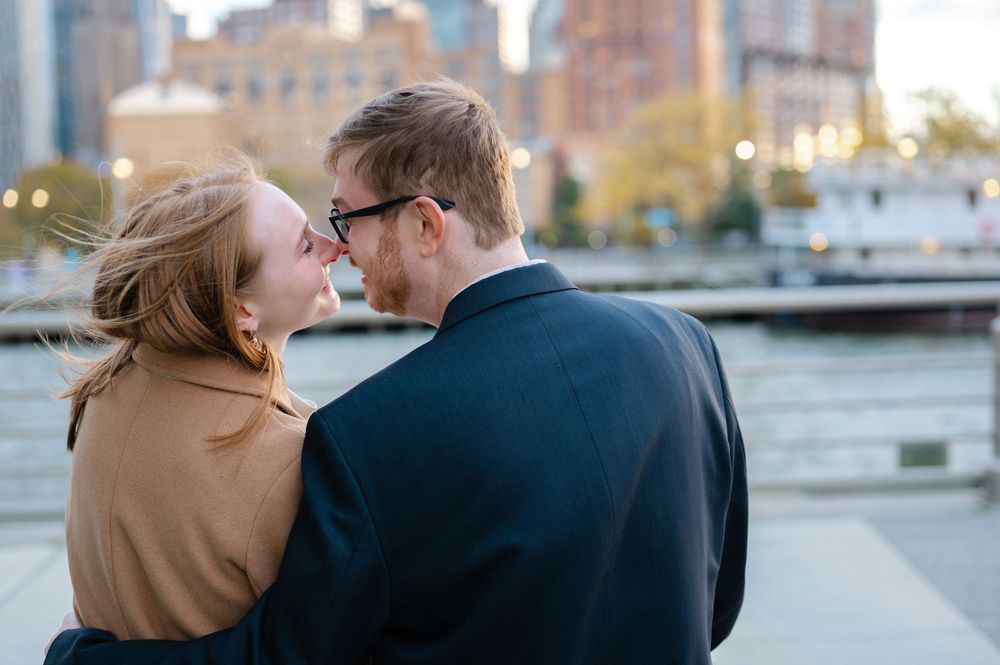 Romantic couple nearly kissing by a waterfront with blurred city buildings behind them.