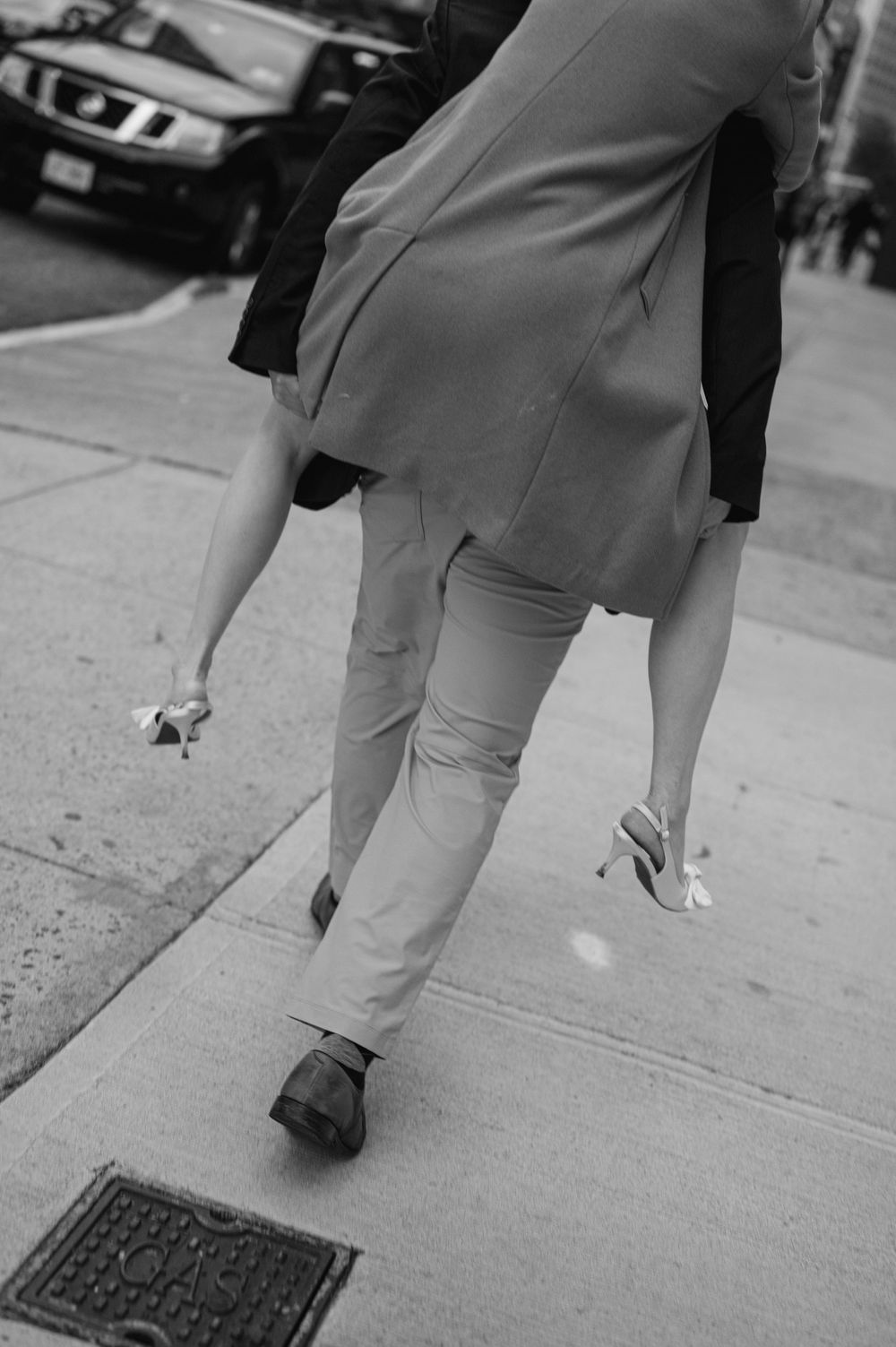 Black and white photo of a couple walking on a city sidewalk, woman carrying heels in hand.