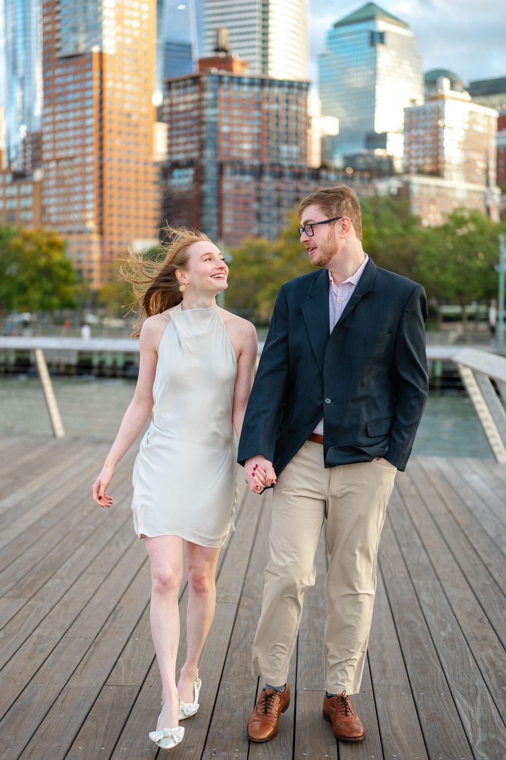 A happy couple walks hand in hand on a waterfront boardwalk with the New York City skyline in the background.