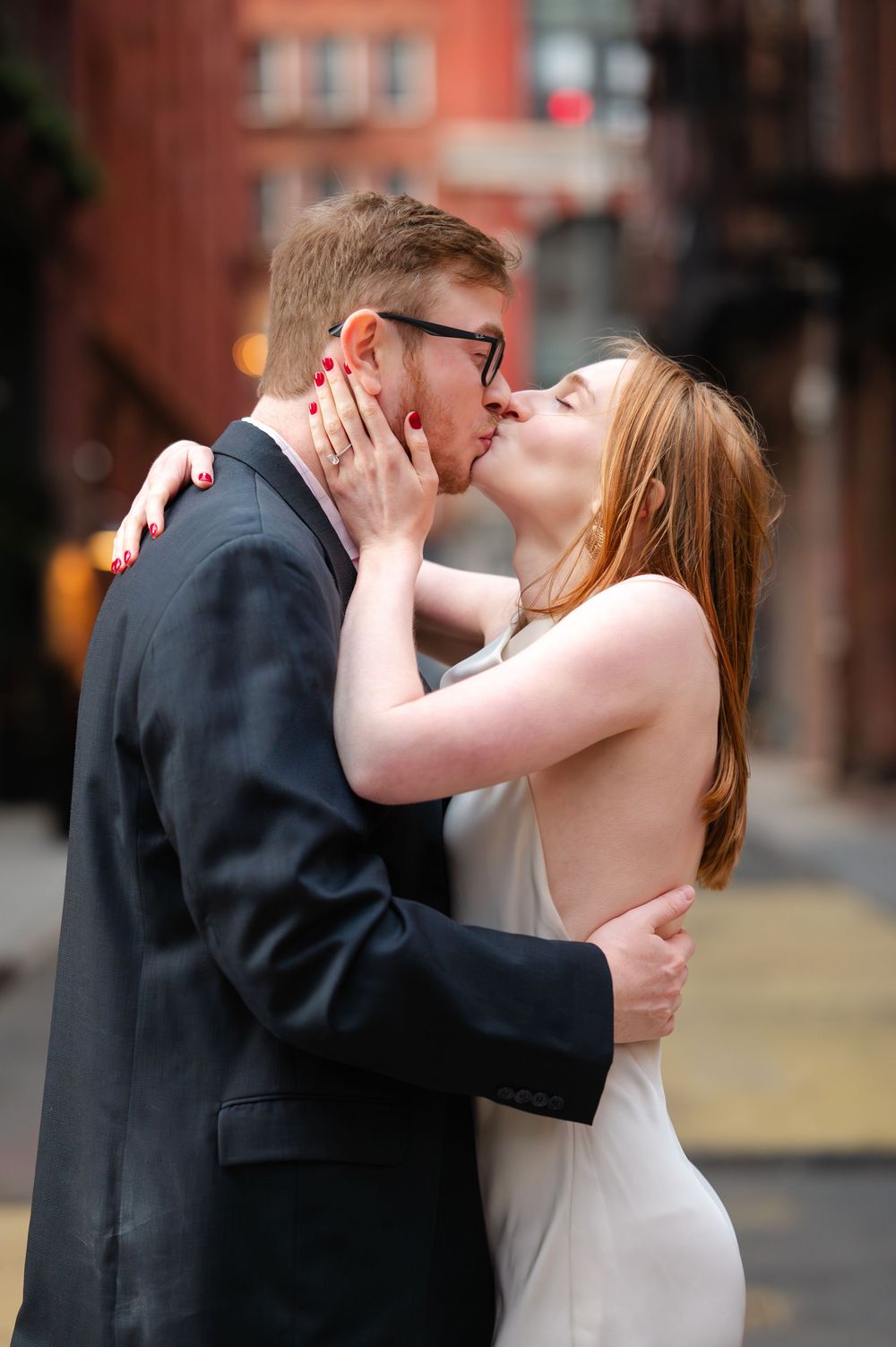 A couple shares a romantic kiss on a city street, the woman in a white dress holding the mans face tenderly.