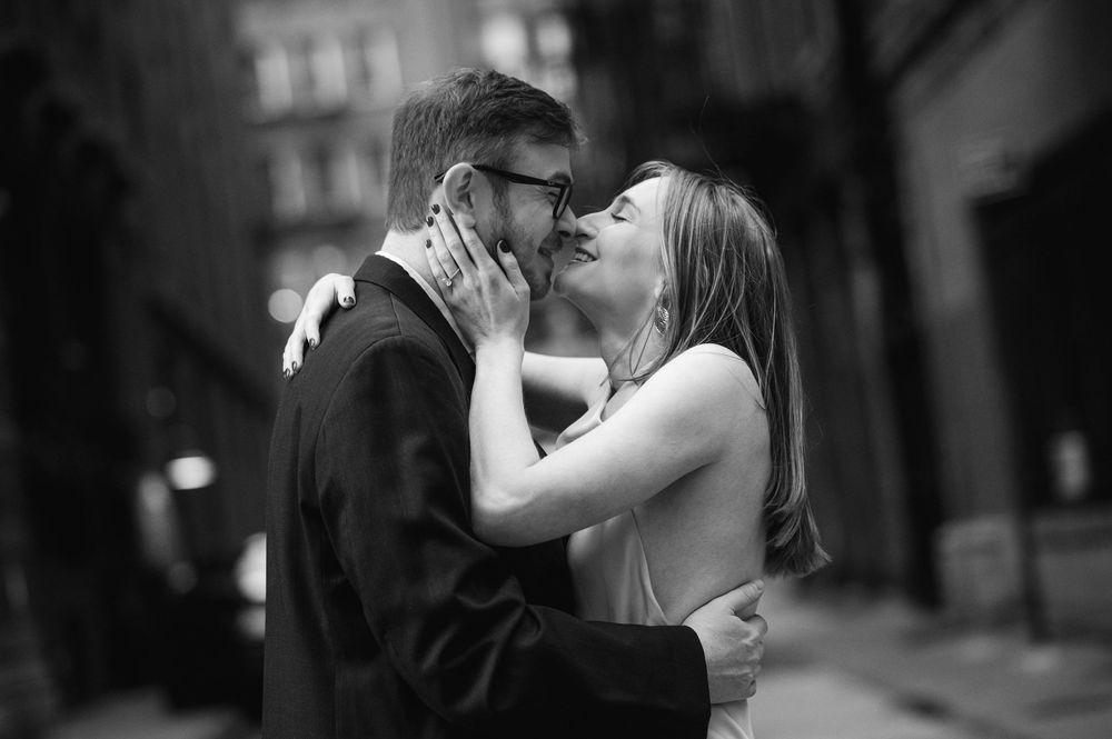 Black and white photo of a couple sharing a romantic kiss on a city street in the rain.