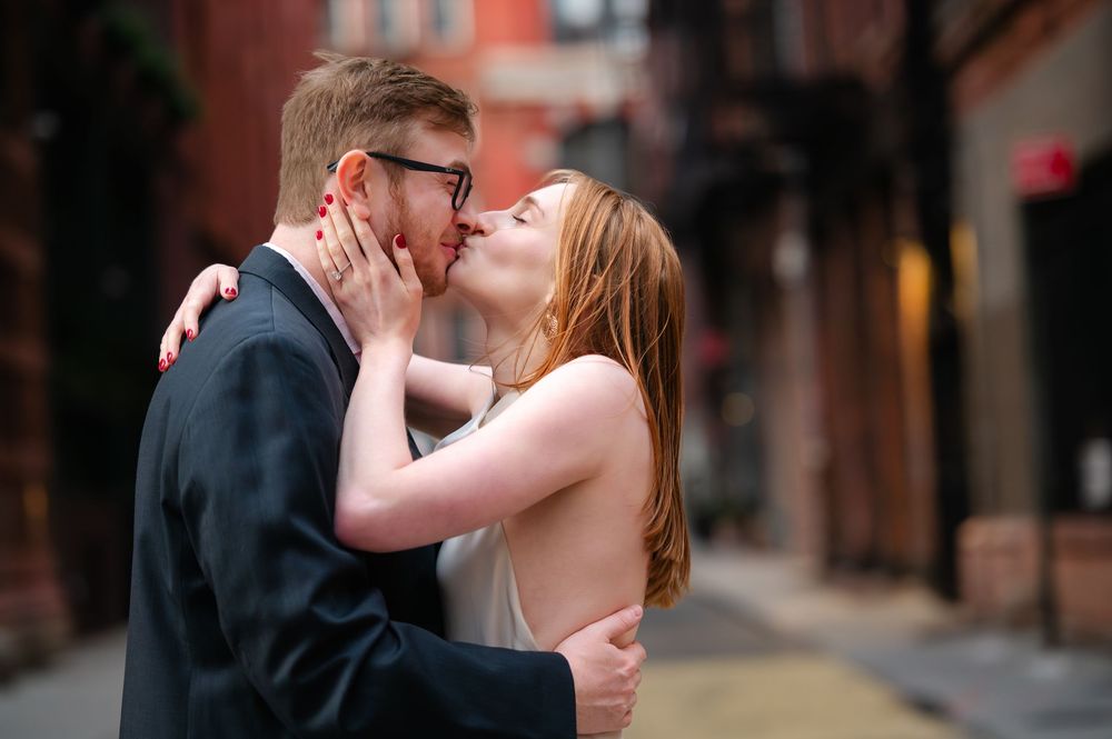 A couple kisses passionately in an urban alley, the woman in a backless white dress embracing the man in a suit.