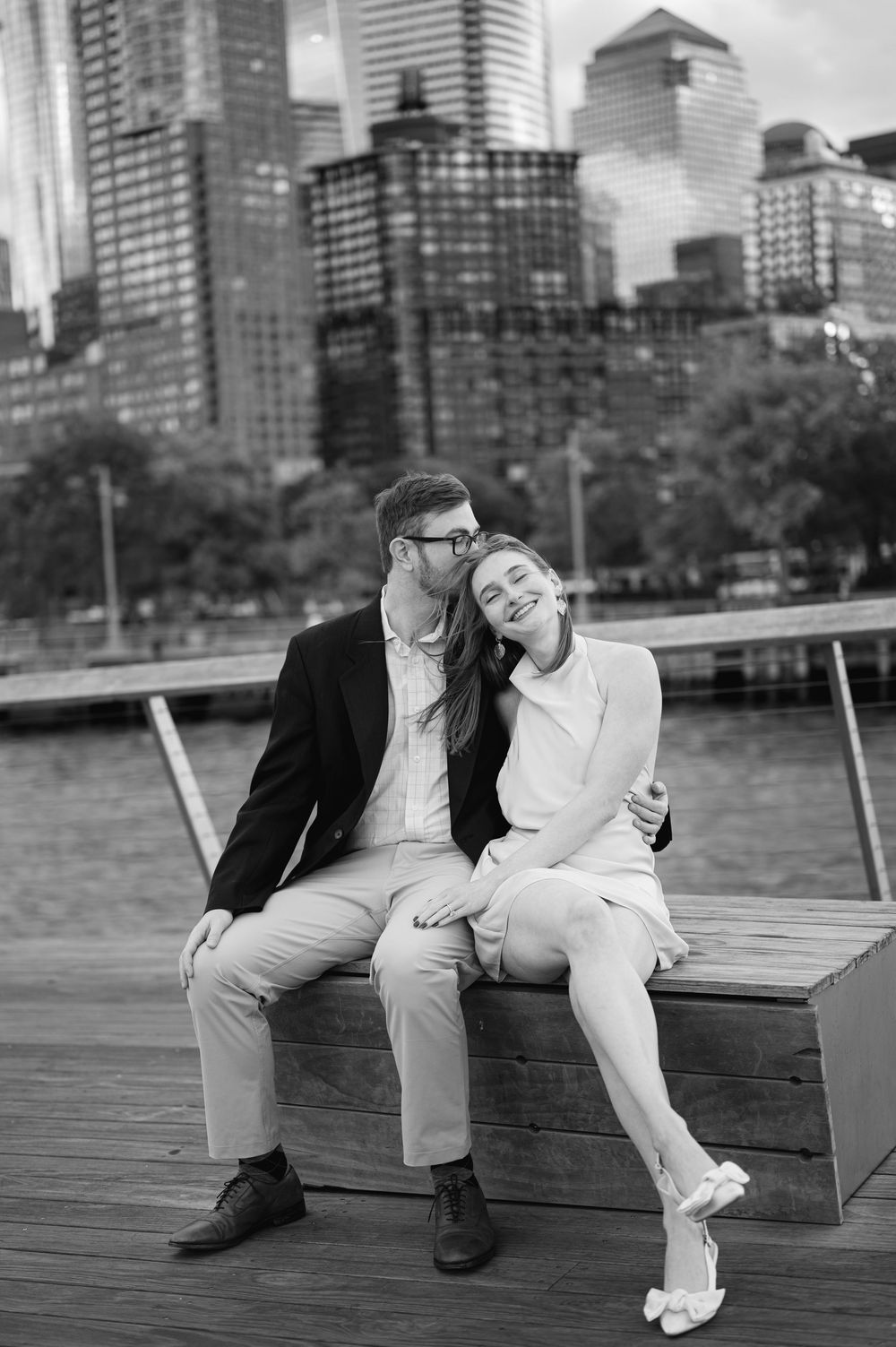 Couple sitting on waterfront bench with NYC skyline, man kissing woman's cheek, black and white engagement photo.
