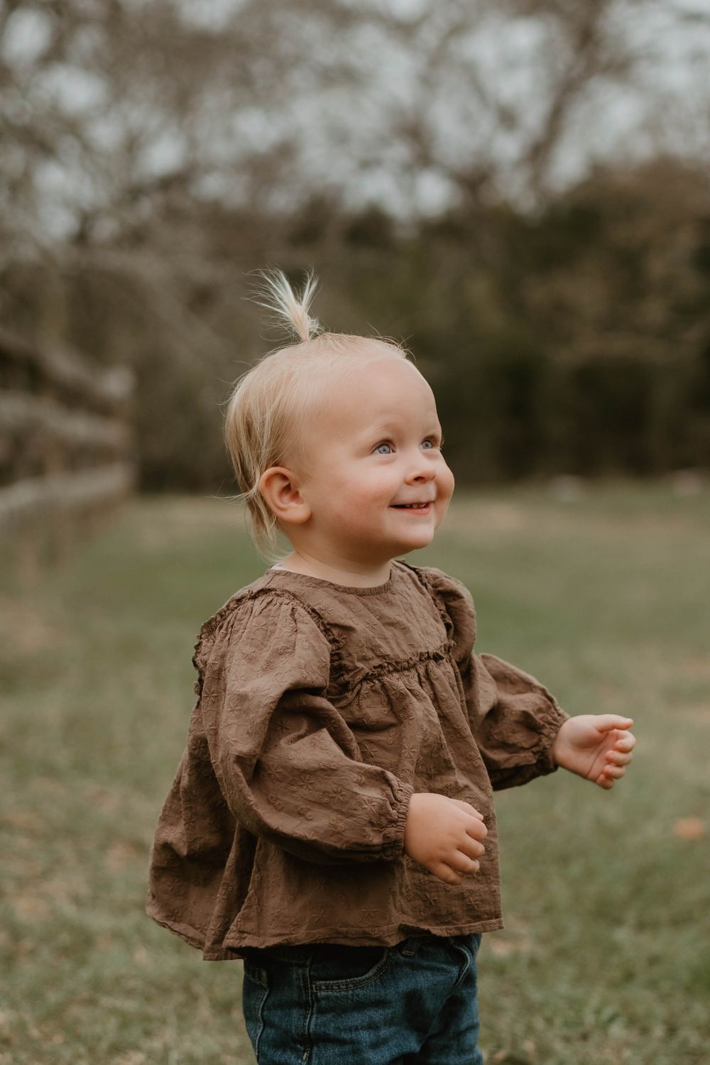 A happy blonde toddler girl in a brown top smiles while standing on green grass outdoors.