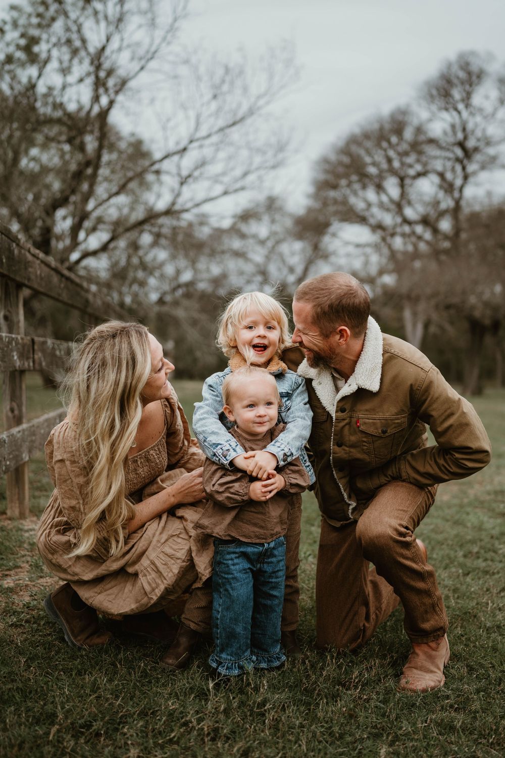 Joyful family with two young children laughing together by a rustic wooden fence in an outdoor photo session.