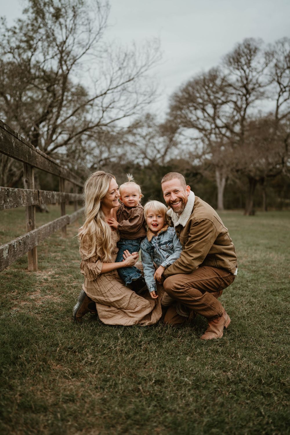 Happy family of four posing outdoors near a wooden fence on a grassy field during a fall family photo session.