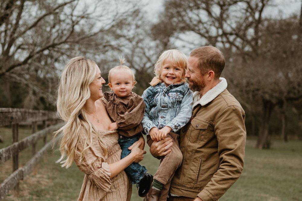 Happy young family with two toddler boys laughing together outdoors in a rustic park setting.