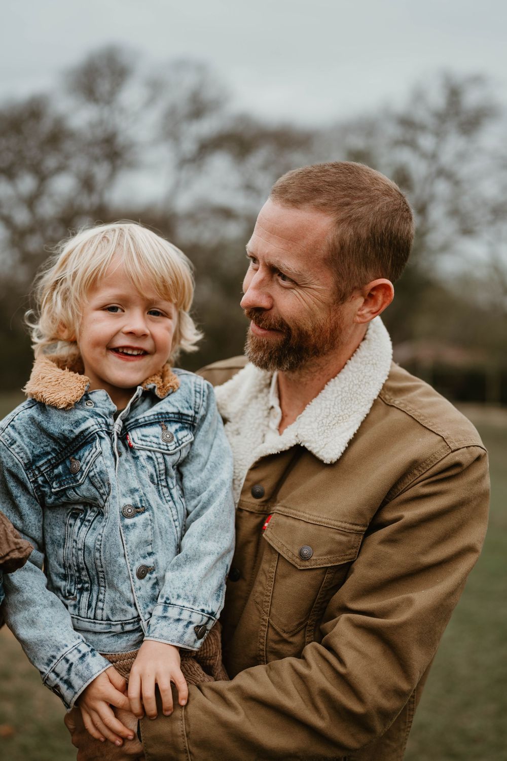 A bearded father holds his laughing blonde toddler son in a denim jacket in a park.