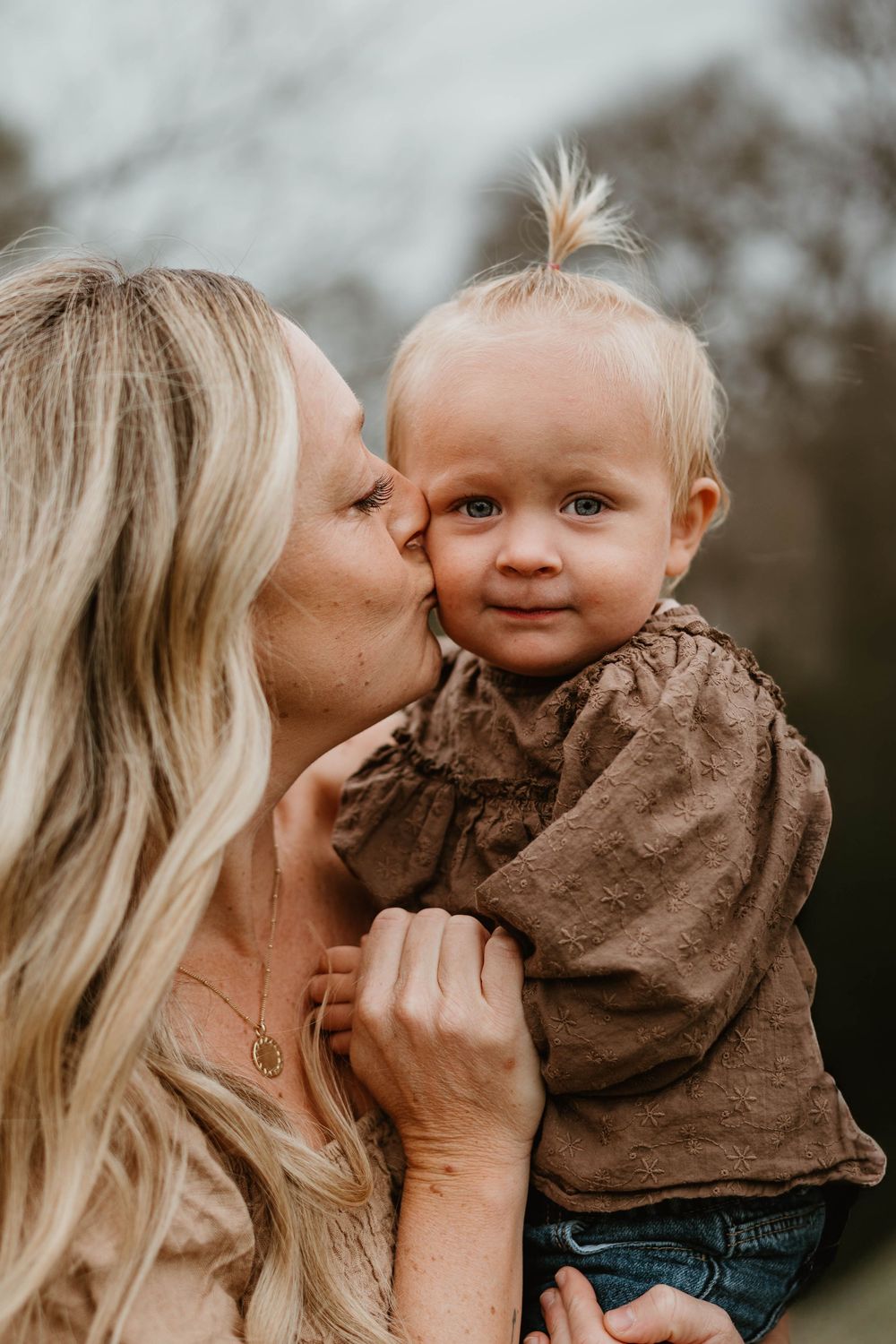 A blonde mother kisses the cheek of her smiling baby girl in a brown top outdoors.