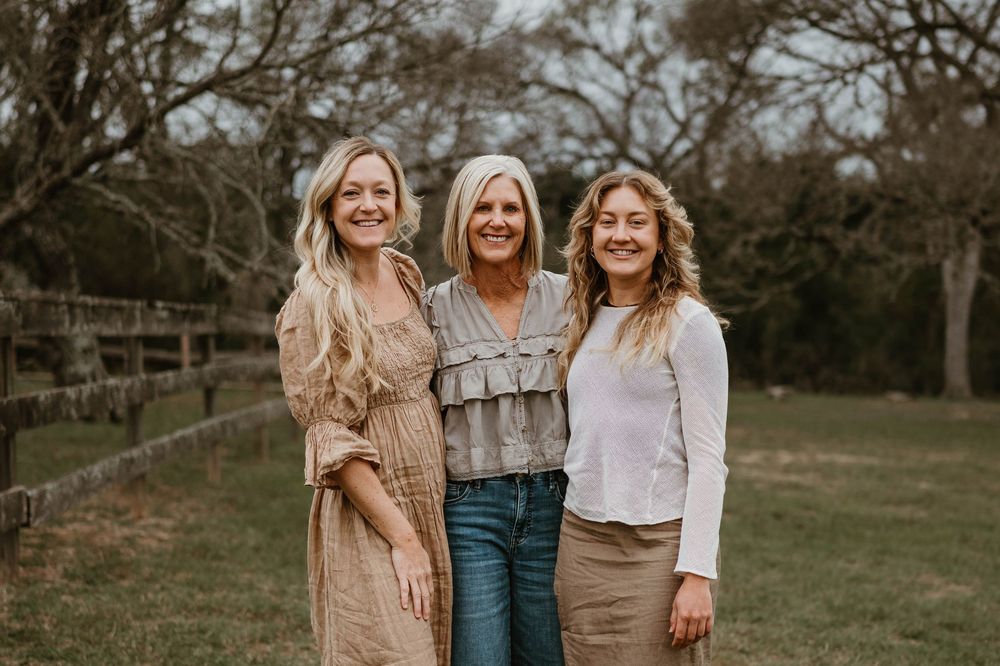 Three smiling women posing together outdoors near a wooden fence during a family portrait session.