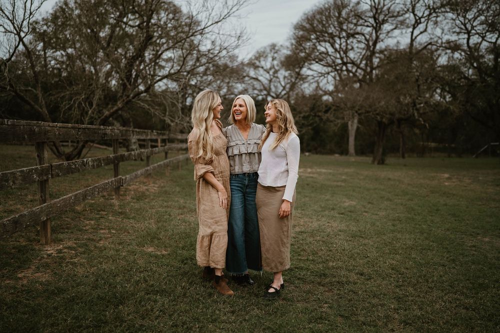 Three blonde women laugh together outdoors on a green lawn near a wooden fence surrounded by bare trees.