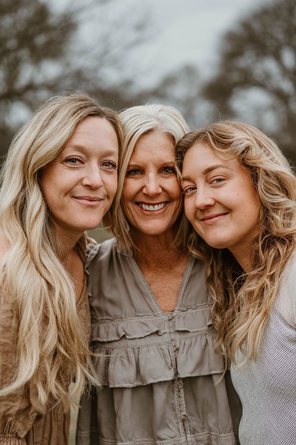 Three blonde women smiling closely together outdoors, likely a mother and two daughters in a warm family portrait.