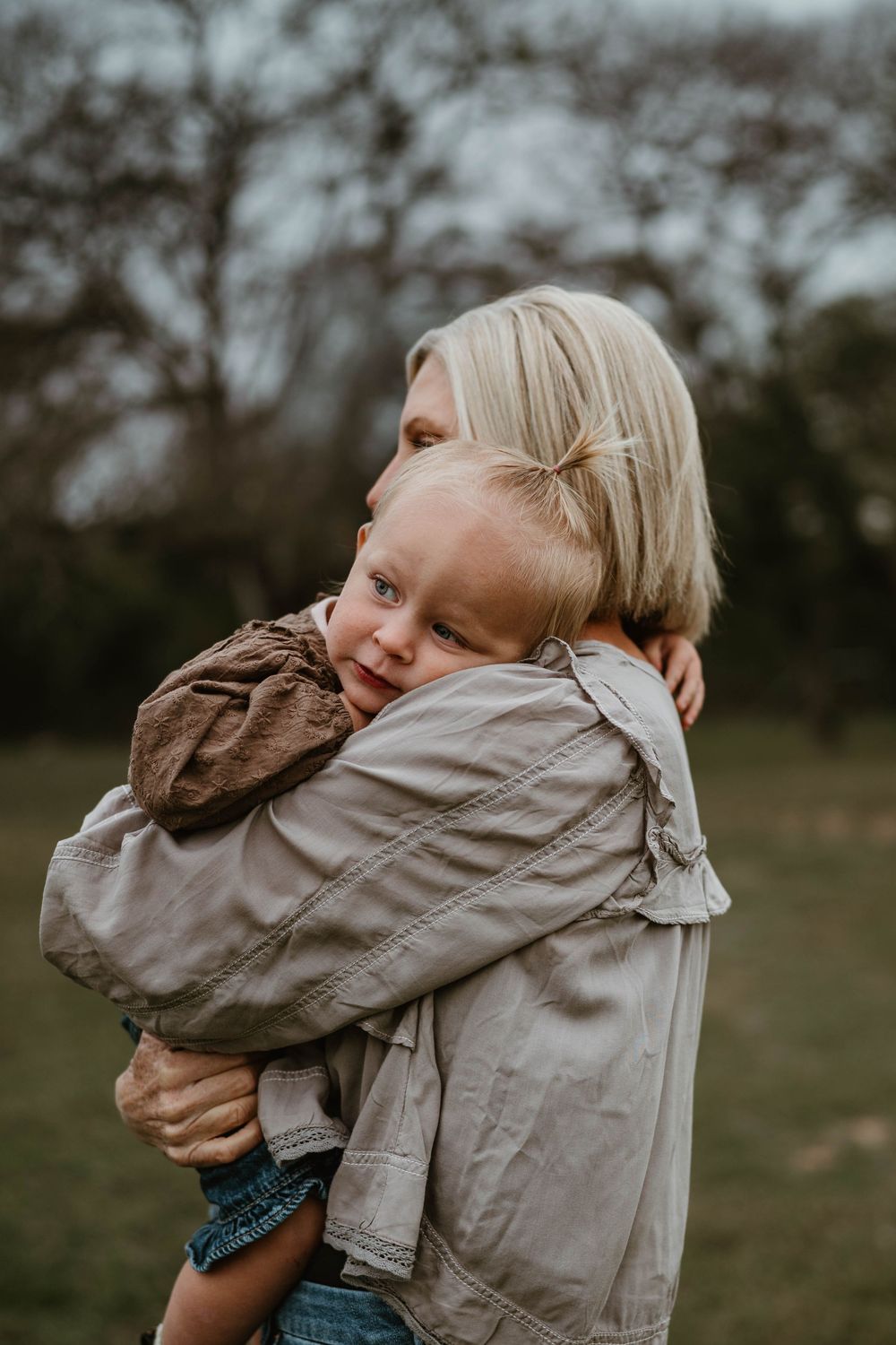 A blonde toddler hugs a woman tightly outdoors in a green park setting.
