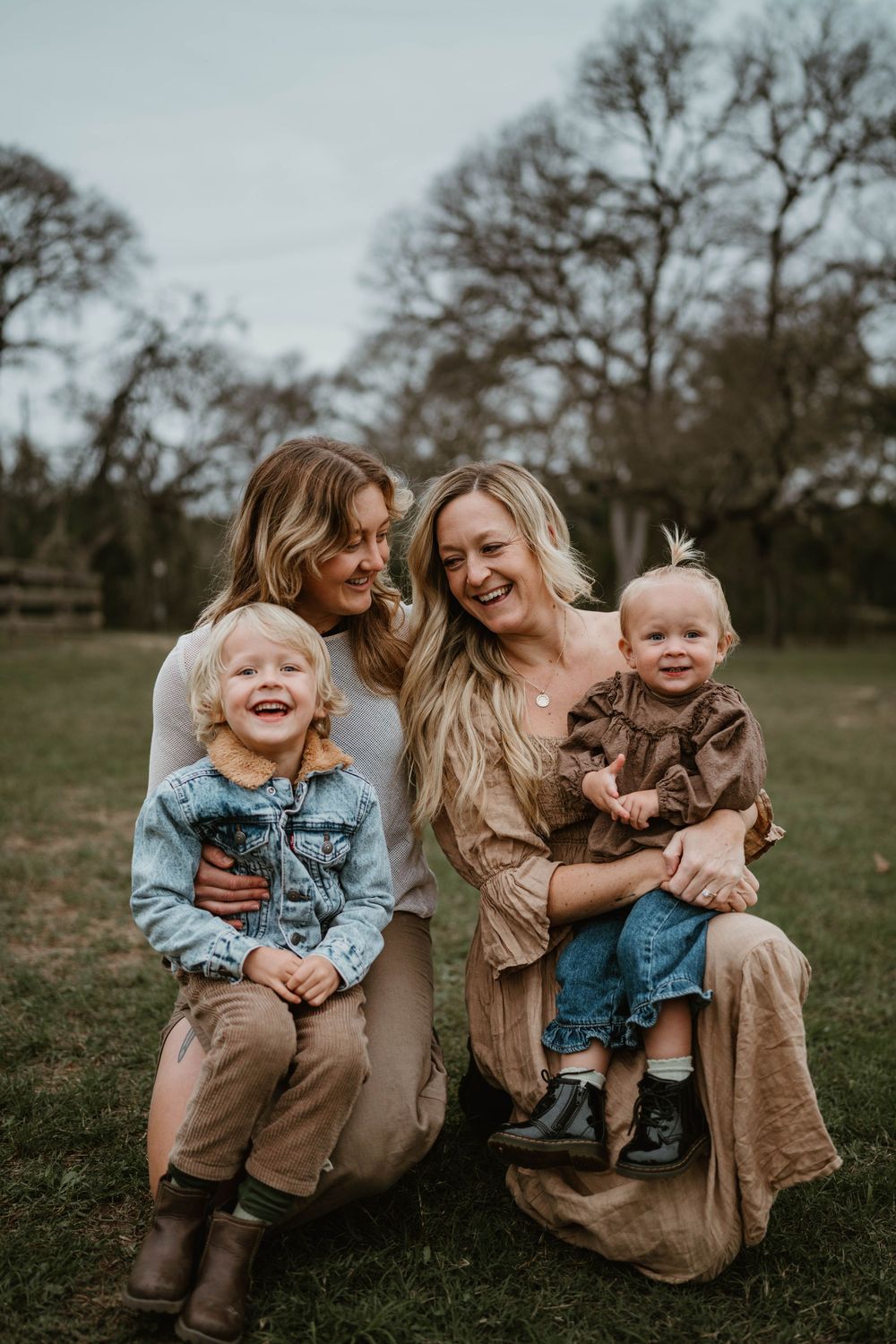Two women smile with two young blonde children on a grassy field on a cloudy day.