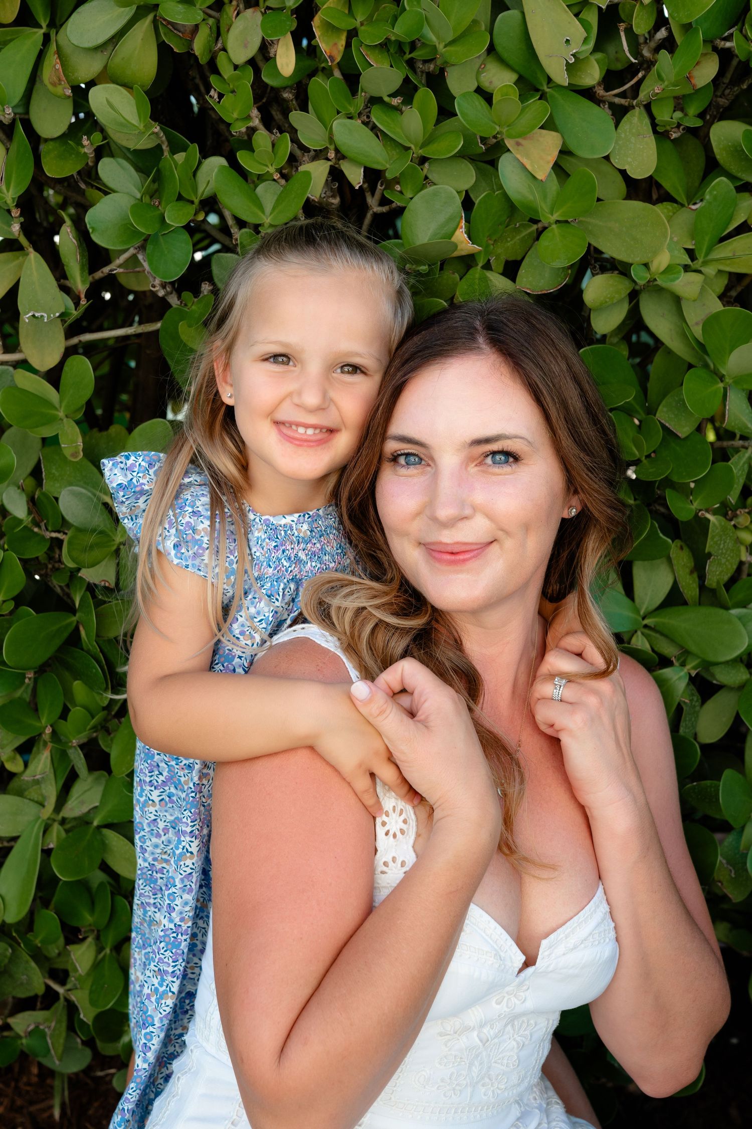 A smiling woman and young blonde girl pose together in front of lush green foliage in matching white and blue outfits.