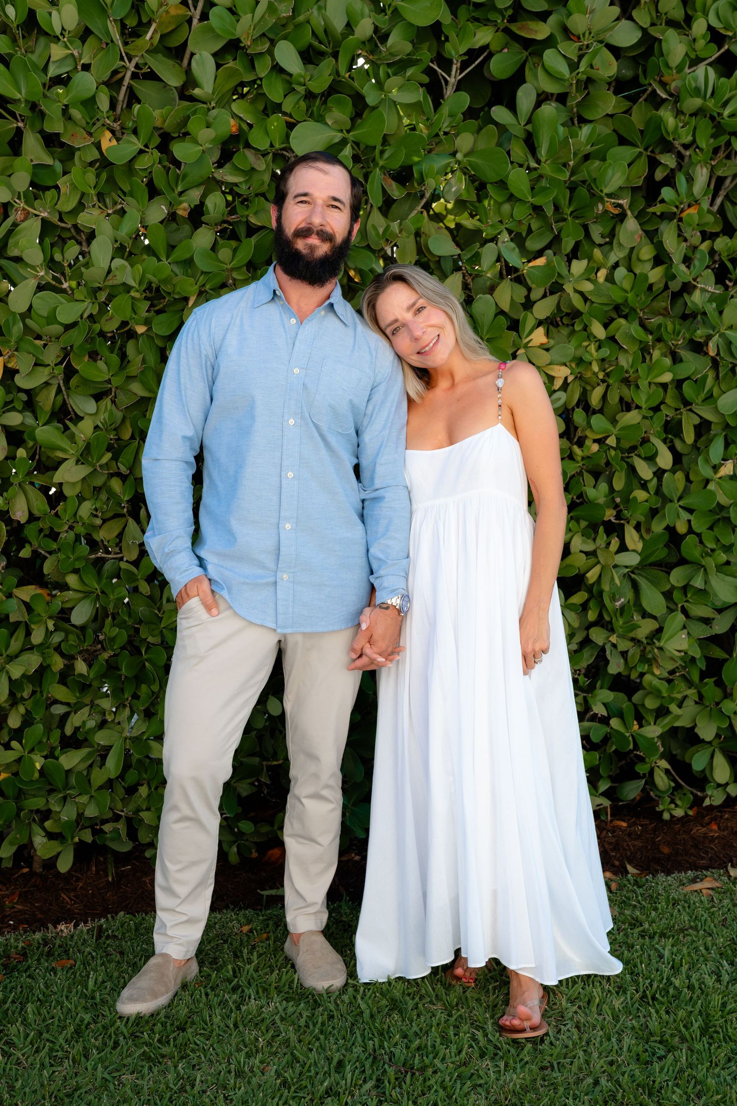 Couple holding hands smiling in front of lush green hedge, man in blue shirt and woman in white dress.