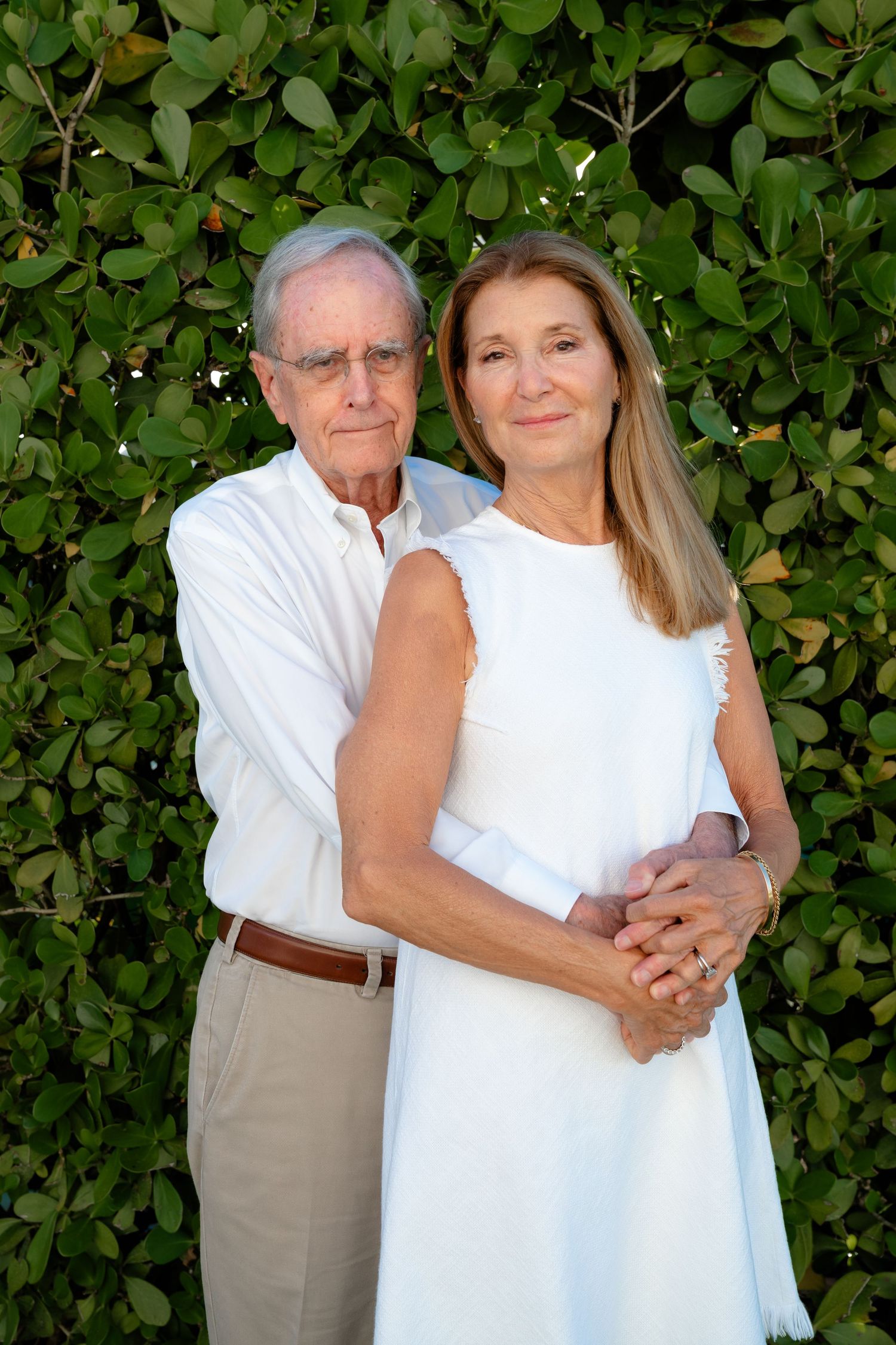 Elegant senior couple in white clothing embracing outdoors against a lush green hedge backdrop.