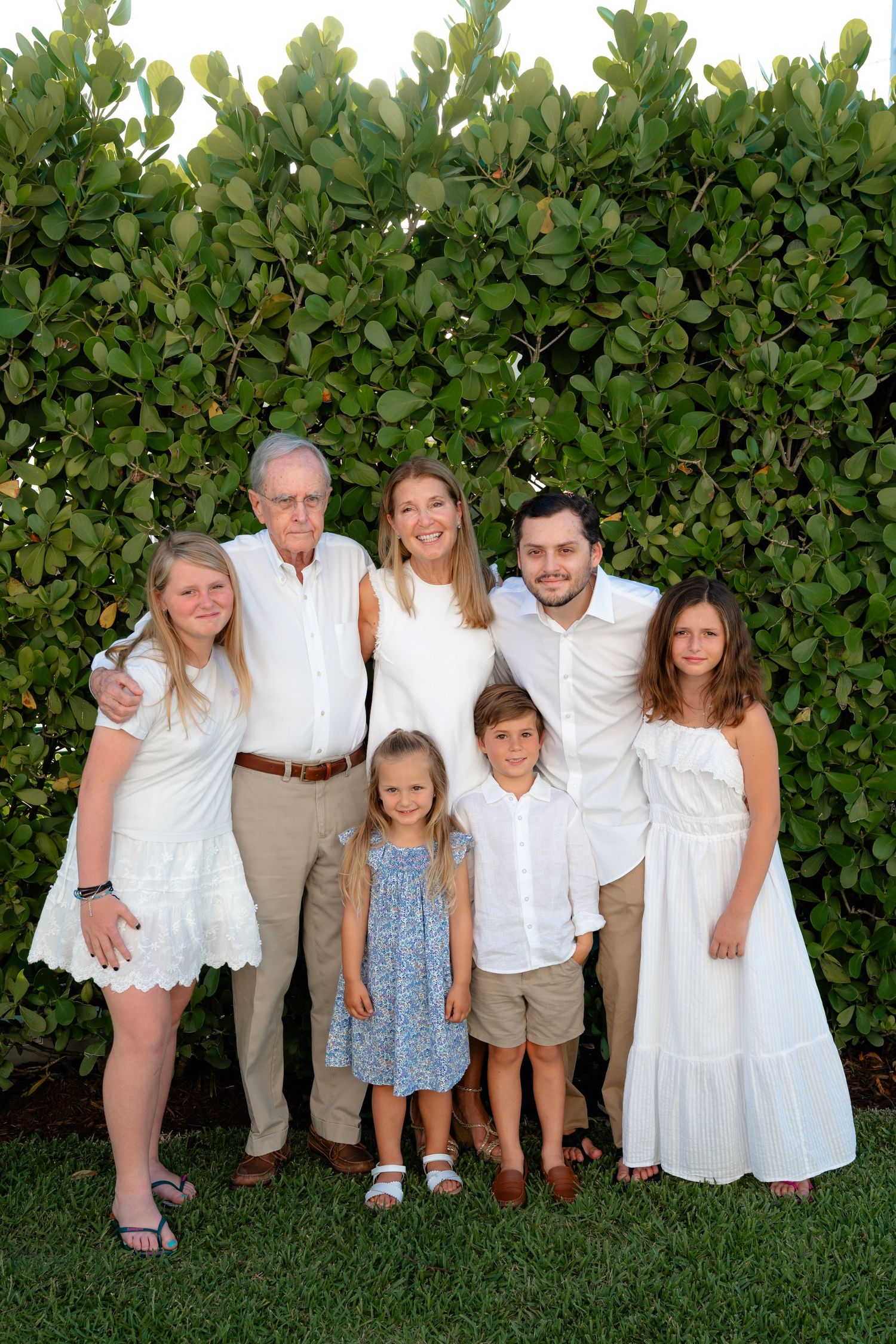 Multi-generational family of seven poses in white outfits against lush green hedge for outdoor portrait photo.