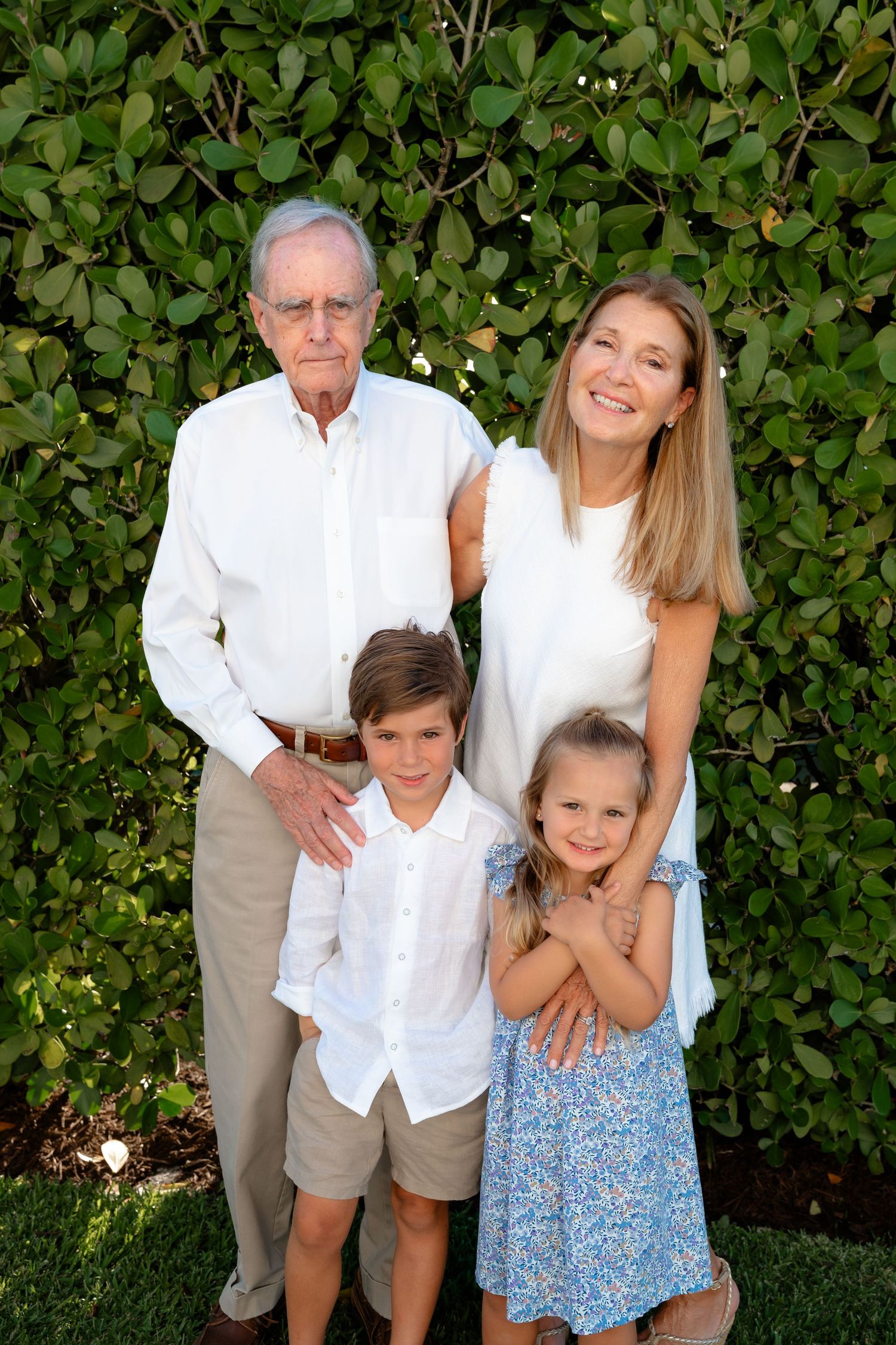 Family portrait of grandpa, mom, and two young children posing outdoors in white outfits before lush green hedge.